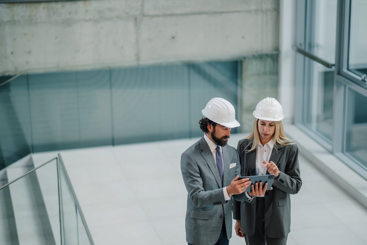 Two architects wearing hard hats reviewing blueprints on a tablet in a modern building